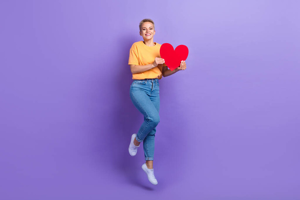 Full length photo of cheerful pleasant girl dressed jeans white sneakers jumping hold red paper heart isolated on violet color background. - Фото, изображение