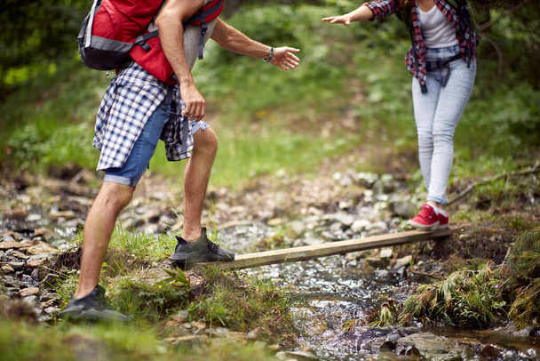 A young couple is helping each other to cross the creek while they hiking on a beautiful day in the nature. Hiking, relationship, nature, activity - Φωτογραφία, εικόνα