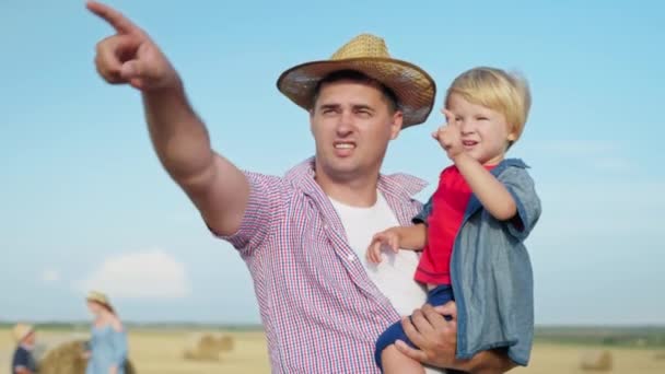父親の世話をする男の親は腕に小さな男の子を抱えていて暖かい夏の日の後に畑で家族の休暇中に干し草で指を指す - 映像、動画
