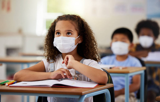 School student with covid learning in class, wearing mask to protect from virus and looking concentrated on education in classroom. Little girl sitting at desk, studying and listening during pandemic. - 写真・画像
