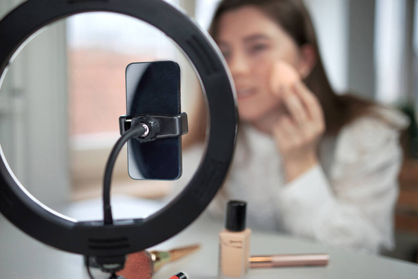 beauty blogging, technology and people concept portrait of a happy smiling girl blogger with ring light and smartphone applying make up at home. making a influencer video Using brush - Photo, image
