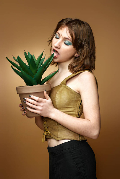 young woman with stylish hairstyle posing with plant in pot on beige background. - Photo, image