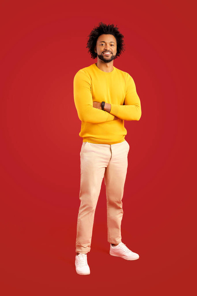 Proud and pleased African-American man in casual wear stands with arms crossed isolated on red, small business owner, male entrepreneur standing in confident pose and looking at camera, hands folded - Фото, изображение