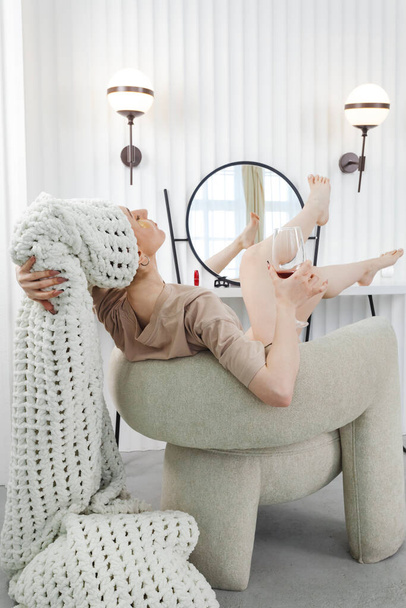 Μια όμορφη νεαρή γυναίκα κάθεται χαλαρή σε μια καρέκλα μετά από ένα ντους με μπαλώματα στα μάτια και μια πετσέτα τυλιγμένη γύρω από το κεφάλι της, κρατώντας ένα ποτήρι κρασί. έννοια της χαλάρωσης και της αυτοφροντίδας. - Φωτογραφία, εικόνα