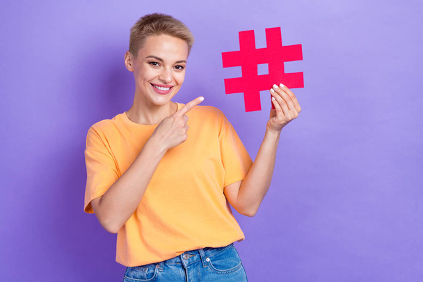 Portrait of positive woman with blond hairstyle wear orange t-shirt directing at red paper hashtag isolated on violet color background. - Foto, Bild