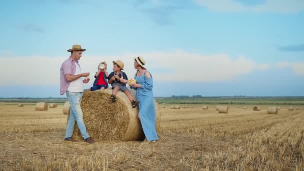 family picnic, happy man and woman with cute boys enjoy spending time together and eat bread washed down with milk from bottle, while relaxing on field with stacks of hay blue sky background - Footage, Video