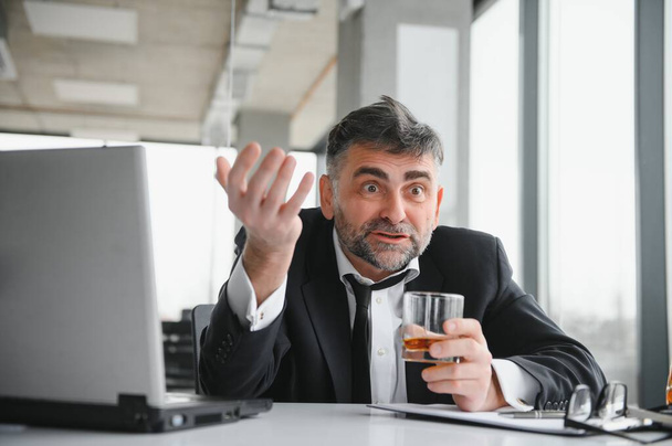 Oude mannelijke werknemer drinkt alcohol op het werk. - Foto, afbeelding