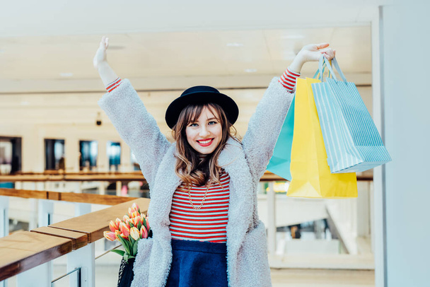 Happy, excited young fashion woman with raised hands holding paper shopping bags on mall background. Woman enjoying new spring season shopping. Consumerism, shopping, sale concept. Selective focus - Fotó, kép