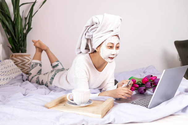 Woman wearing facial mask enjoying at home and using her laptop. young Asian woman with beauty treatment facial mask using credit card online shopping on bed. - Foto, imagen