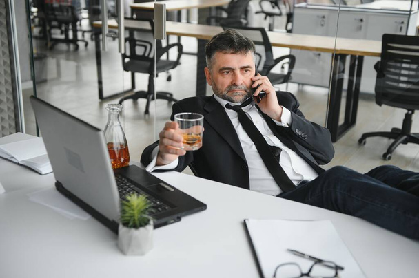 Old male employee drinking alcohol at workplace. - Zdjęcie, obraz