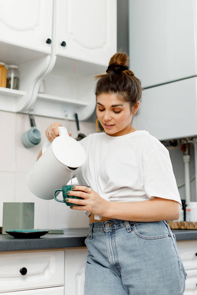 Uma menina morena com cabelo estilizado em um pão está fazendo chá na cozinha pela manhã. Uma menina em uma camiseta branca e jeans está fazendo chá em uma cozinha branca. - Foto, Imagem