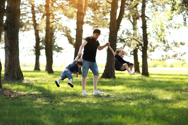Ημέρα πατέρων. Ευτυχισμένος μπαμπάς διασκεδάζει και γυρίζει δύο παιδιά έξω. Ελεύθερος μπαμπάς με μικρό γιο και κόρη χαλαρώνοντας στο καλοκαιρινό πάρκο. Οικογενειακή μέρα και παιδική ηλικία. Ενεργό αστείο παιχνίδια, ζωντανό παιχνίδι. - Φωτογραφία, εικόνα
