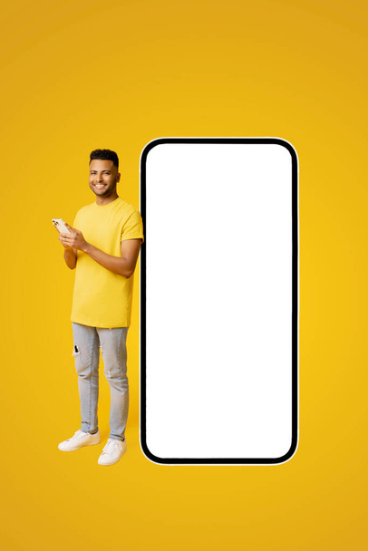 Handsome happy indian guy standing near big huge smartphone with blank screen, copy space, mockup, full length latin man in yellow t-shirt using mobile phone, texting near big phone - Фото, изображение