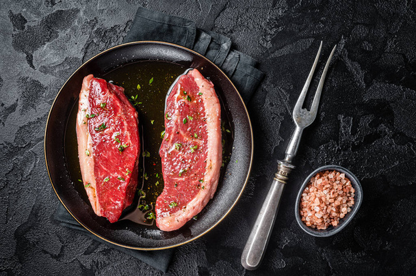 Raw marbled beef meat steak marinated with olive oil, thyme, salt and pepper. Black background. Top view. - Photo, Image