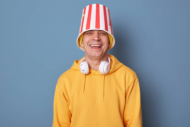 Portrait of happy overjoyed man wearing beanie hat and casual yellow hoodie posing against blue wall, standing with popcorn bucket on head, laughing out loud, having fun in cinema - Fotoğraf, Görsel