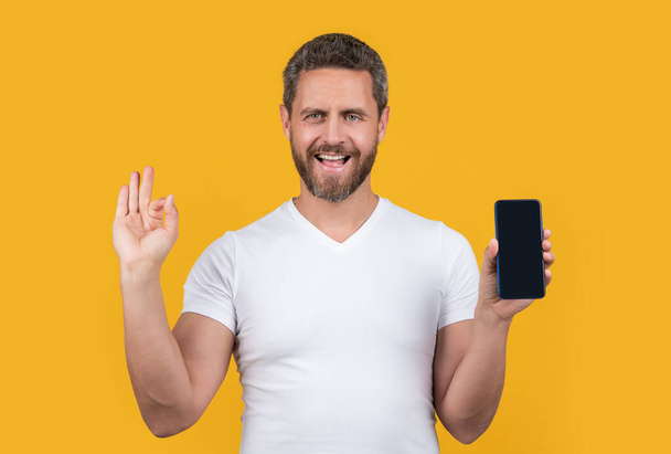 glad man showing phone screen with copy space. man showing phone screen isolated on yellow background. man showing phone screen in studio. man showing screen of phone. - Photo, Image