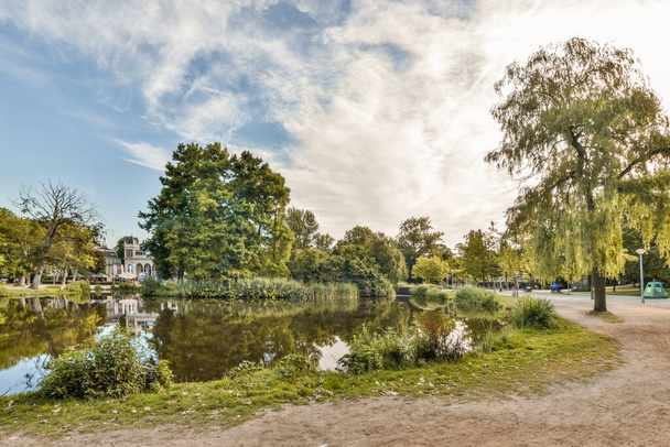 Amsterdam, Netherlands - 10 April, 2021: a lake in the middle of a park with trees and people walking on the path to the waters edge - Fotó, kép