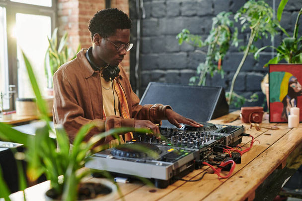 jovem afro-americano transformando misturadores e gira-discos de equipamentos musicais modernos, enquanto em pé à mesa de madeira longa - Foto, Imagem