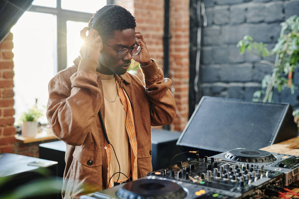 Joven hombre negro poniendo auriculares en su cabeza mientras está de pie junto a la mesa con equipo musical en la sala de estar del apartamento loft - Foto, imagen