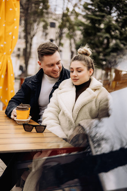 Glückliches junges verliebtes Paar in Oberbekleidung, das draußen an einem Tisch sitzt und Kaffee trinkt. Entspannen an der frischen Luft - Foto, Bild