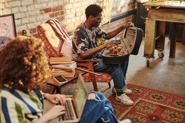Young African American man opening case with violin while sitting in armchair in front of his girlfriend with laptop on her knees - Photo, Image