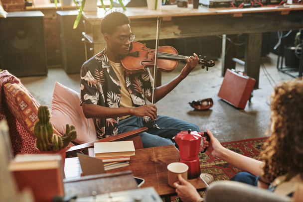 Joven negro con instrumento musical de cuerda interpretando pieza de música clásica frente a su novia sosteniendo taza y tetera - Foto, Imagen