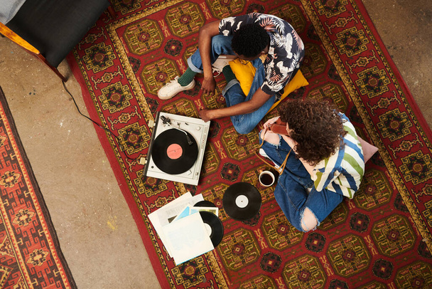 Верхній кут двох зумів у стильному повсякденному одязі, що слухає диски на рекордному плеєрі, розслабляючись на червоному килимі у вітальні
 - Фото, зображення