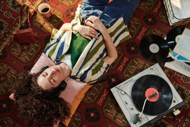 Nad úhlem mladé ženy v casualwear těší hudbu ve sluchátkách, zatímco odpočívá na červeném koberci na podlaze - Fotografie, Obrázek