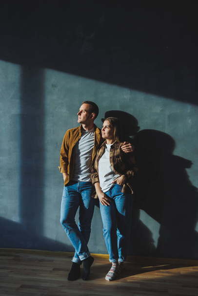 A young married couple in love in shirts and jeans on the background of a gray wall. The concept of happy family relationships. A man and a woman are hugging - Zdjęcie, obraz