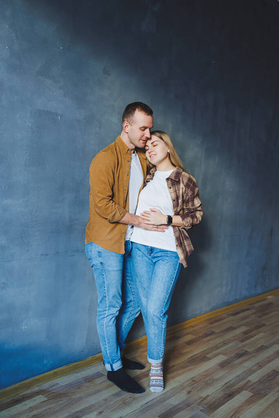 A young married couple in love in shirts and jeans on the background of a gray wall. The concept of happy family relationships. A man and a woman are hugging - Foto, Imagen