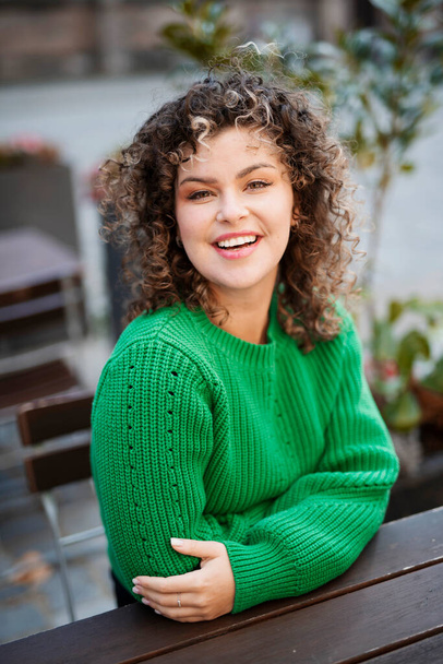 Joven mujer sonriente en un suéter verde sentado en la cafetería, concepto de moda callejera - Foto, Imagen