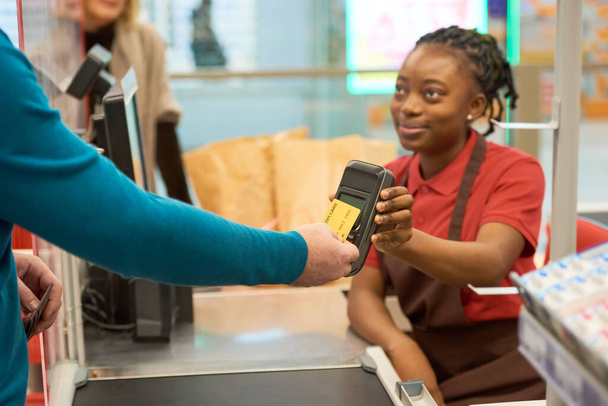 Modern alıcı seçilmiş malları kredi kartıyla öderken aynı zamanda genç Afrikalı Amerikalı kadın satıcının elinde tutuyor. - Fotoğraf, Görsel