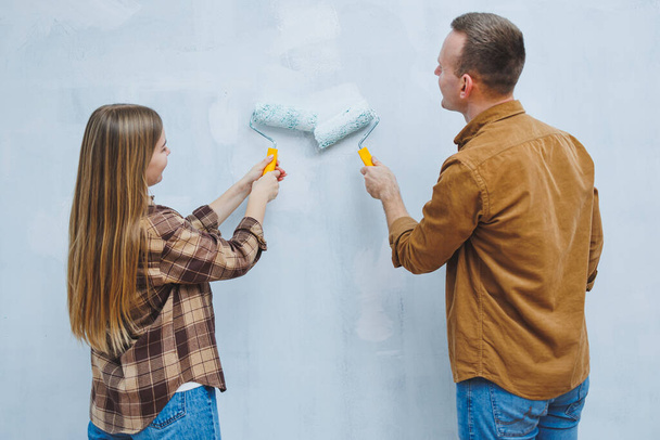 Couple in love, newlyweds in shirts and jeans in an empty room, painting white walls and renovating the interior. A man and a woman moved to a new house. Selective focus - Photo, Image