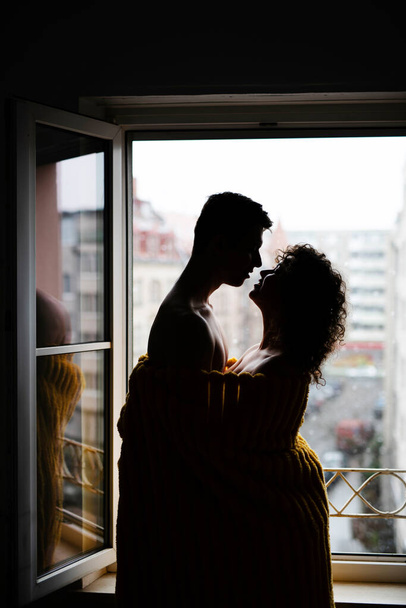 A young couple hugging in a blanket, photography against the light, a couple in love near the window in a blanket - Photo, Image