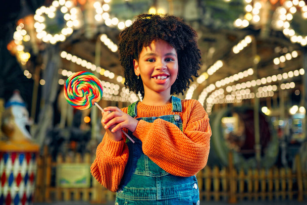 アフロの髪型のかわいいアフリカ系アメリカ人の女の子は、遊園地やサーカスで夜に馬とカルーセルの背景にカラフルなロリポップを食べます. - 写真・画像