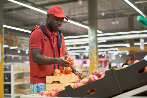 Schwarzer Mann in roter Uniform putzt frische reife Äpfel aus einer Holzkiste zusammen mit anderem Obst und Gemüse in der Lebensmittelabteilung - Foto, Bild