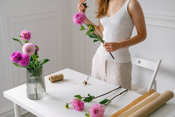 Εύθραυστη γυναίκα ανθοπώλης συλλέγει μπουκέτο από ροζ ντάλιες σε ένα βάζο στο λευκό τραπέζι. Μια εικόνα για το λουλουδάτο σχέδιό σου. Η έννοια της άφιξης της άνοιξης - Φωτογραφία, εικόνα