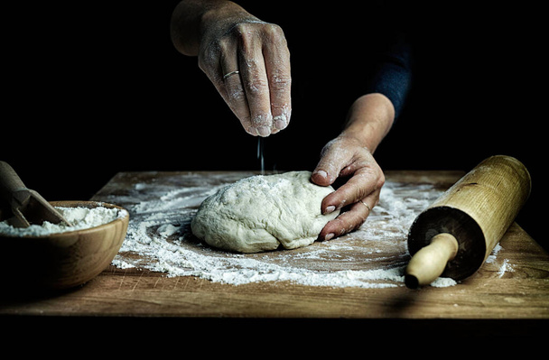Het bereiden van traditioneel zelfgemaakt pasta deeg. Close-up zicht op de handen van de vrouw kneden vers deeg voor het maken van brood of pizza op een bloemrijke tafel. Donkere achtergrond. Kopieerruimte. - Foto, afbeelding