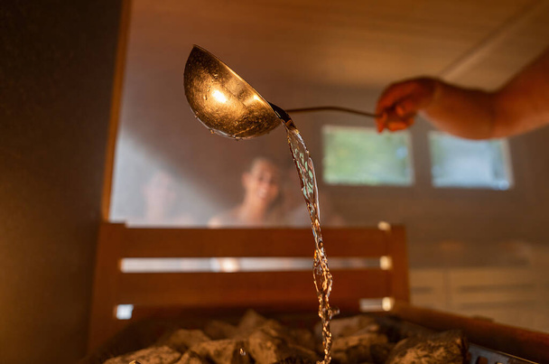 Bir adam sauna odasında bir grup insanla sıcak taşın üzerine metal bir spo döktü. Taşlarda su buharı, spa ve sağlık konsepti, sıcak finiş saunasında rahatlamak. Sıcak sıcak banyo terapisi.. - Fotoğraf, Görsel