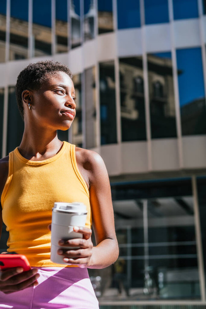 通りで待っている非常に短い髪を持つ若いと魅力的なアフリカの女性。彼女は再利用可能なコーヒーマグカップとスマートフォンを持っています. - 写真・画像