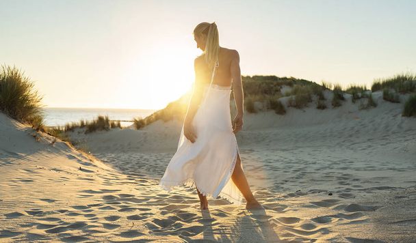 Frau steht barfuß durch Sanddünen zum Meer bei sommerlichem Sonnenuntergang - Foto, Bild