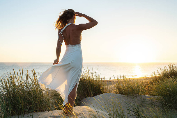 jovem mulher atraente standin em dunas pela praia do mar olhando para o oceano ao pôr do sol - Foto, Imagem