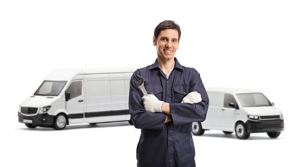Young worker in a uniform holding a wrench in front of two white vans isolated on white background - Valokuva, kuva