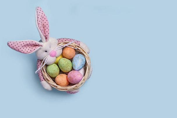 Una cesta de Pascua festiva llena de coloridos huevos decorados y un lindo conejito de felpa en un fondo azul pastel minimalista perfecto para diseños y decoraciones con temática navideña. - Foto, imagen
