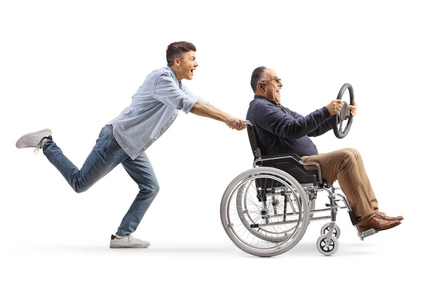 Full length profile shot of a guy pushing a mature man in a wheelchair holding a steering wheel isolated on white background - Foto, immagini