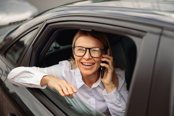 Businesswoman traveling on back seat of car and talking smartphone while looking at window - Фото, изображение