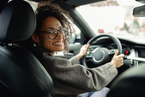 Vrouw rijdt auto en houdt beide handen op het stuur op weg naar het werk en kijkt naar de camera - Foto, afbeelding