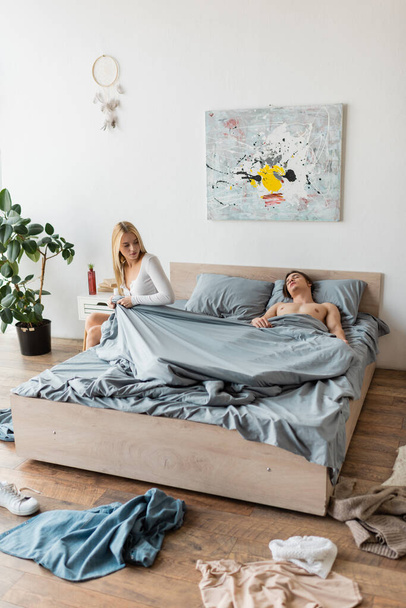 blonde woman sitting on bed and pulling blanket while shirtless man sleeping after one night stand   - Foto, afbeelding