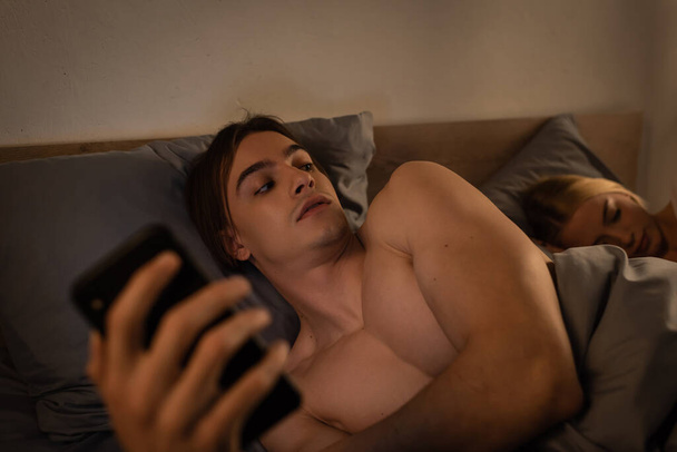 unfaithful man texting on smartphone and looking at blonde girlfriend lying in bed, cheating concept  - Photo, Image