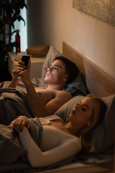 unfaithful man using mobile phone near blonde girlfriend lying in bed, cheating concept  - Photo, Image
